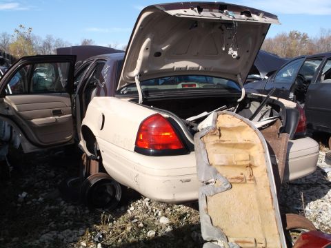 1999 Mercury Grand Marquis - A beige sedan with its trunk and driver's side door open is parked in a junkyard. A torn seat cushion lies in the open trunk.