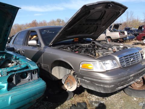 1999 Mercury Grand Marquis - A gray sedan with its hood open is parked in a junkyard, revealing its engine components. Another vehicle, a teal-colored car, is partially…