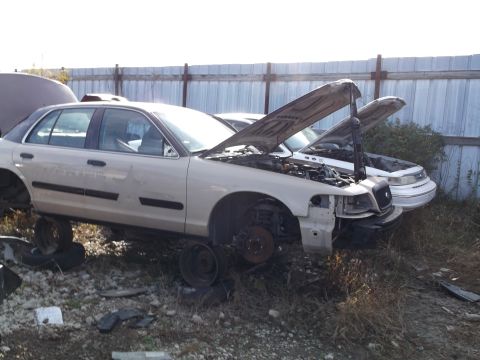1999 Mercury Grand Marquis - Two older model sedans, one tan and one white, are parked in a junkyard with their hoods open. The tan car in the foreground is missing its…