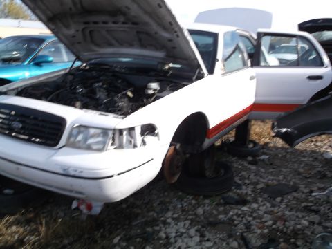 1999 Mercury Grand Marquis - A white sedan with its hood open is parked in a junkyard. The vehicle has an orange stripe along its side and appears to be missing some pa…
