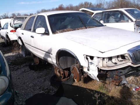 1999 Mercury Grand Marquis - A white sedan, likely a Mercury Grand Marquis, sits in a junkyard with its front end partially dismantled and missing its bumper and headli…