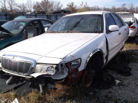 1999 Mercury Grand Marquis - A damaged white Mercury Grand Marquis is parked in a junkyard with its front end heavily damaged. The car is missing its front bumper and h…