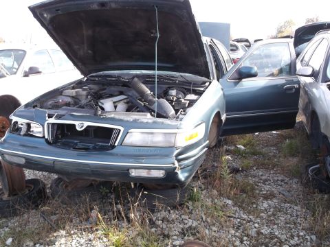 1999 Mercury Grand Marquis - A dark green Mercury Grand Marquis with its hood and driver's side door open sits in a junkyard. The front of the car is visible, showing t…