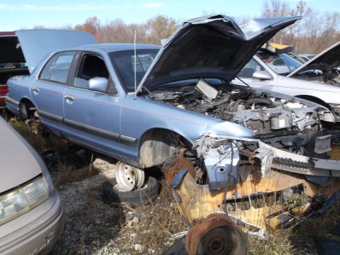 1999 Mercury Grand Marquis - A light blue sedan with its hood open, showing the engine compartment. The vehicle is in a junkyard setting with other vehicles and debris…