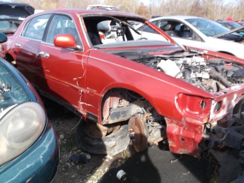 1999 Mercury Grand Marquis - A damaged red sedan is parked in a junkyard with its hood open and engine exposed. Another vehicle is parked closely to its left.