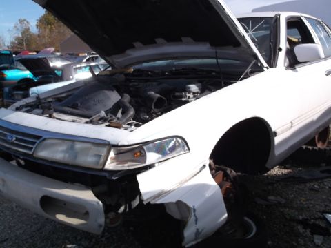 1999 Mercury Grand Marquis - The front end of a white sedan is shown with its hood open, revealing the engine bay. The vehicle appears to be in a junkyard setting with…