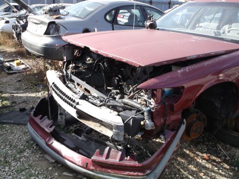 1999 Mercury Grand Marquis - A damaged maroon Mercury Grand Marquis is parked in a junkyard with its hood open, revealing the engine compartment. The front bumper and g…