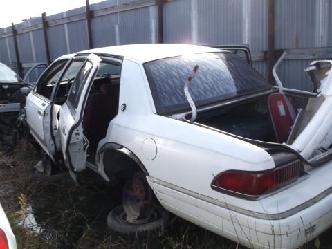 1999 Mercury Grand Marquis - A white 1999 Mercury Grand Marquis is parked in a junkyard with its doors and trunk open. The car is missing its front tires and the rear t…