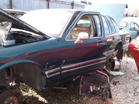 1999 Mercury Grand Marquis - The driver's side of a dark green sedan is shown in a junkyard setting. The hood and driver's door are open, and the front wheel is missing.