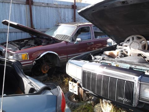 1999 Mercury Grand Marquis - Two older model sedans are parked in a junkyard. The front vehicle has its hood open, revealing the engine and a chrome grille with "LINCOL…
