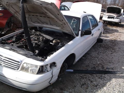 1999 Mercury Grand Marquis - A white sedan with its hood open is parked in a junkyard. The engine bay is visible, and parts of the front end, including the headlight an…