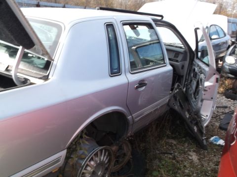 1999 Mercury Grand Marquis - The side and rear of a light silver sedan is shown in a junkyard setting. The driver's side rear door is open and partially disassembled, r…
