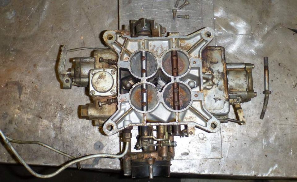 Disassembled four-barrel carburetor with rust on a metal workbench.