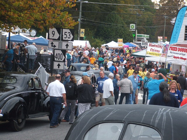 Black vintage coupe at an outdoor festival with crowds and tents.