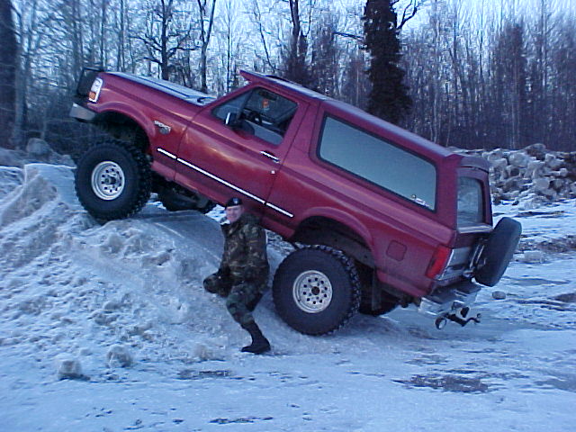 Red lifted Ford Bronco tilted up on a snow bank as a person in camo stands beside the front tire