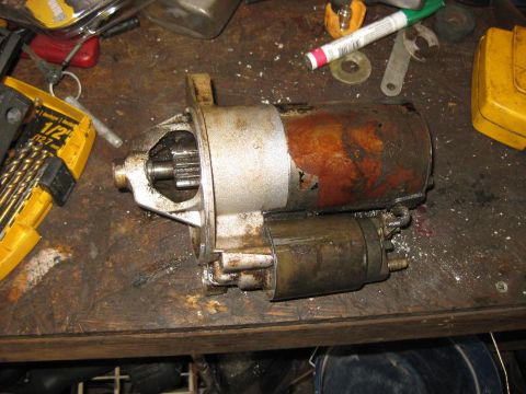 1994 Ford Bronco - A used and rusty starter motor for a 1994 Ford Bronco is shown on a wooden workbench with various tools and parts scattered around.