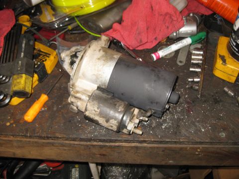 1994 Ford Bronco - A used starter motor is shown on a workbench with various tools and shop rags. The starter motor is dirty and appears to have been recently…