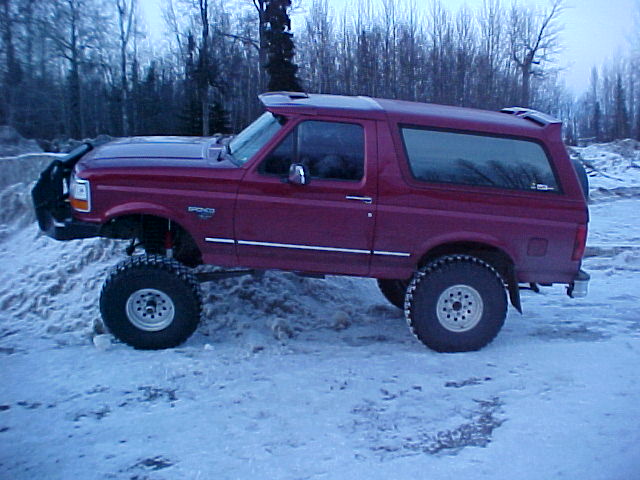 Side profile of red lifted Ford Bronco with tube bumper on snow, wooded ridge behind