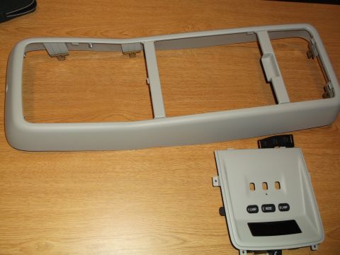 1999 Mercury Grand Marquis - Two gray plastic automotive interior trim pieces are shown on a wooden surface. The larger piece is a rectangular bezel with mounting tabs,…