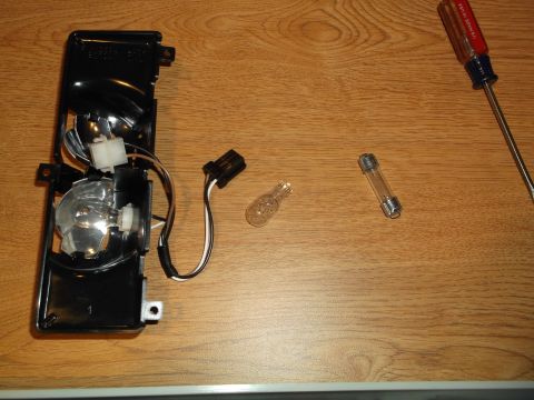1999 Mercury Grand Marquis - A disassembled black automotive light housing with a bulb and wiring is shown on a wooden surface, next to a glass fuse and a screwdriver.