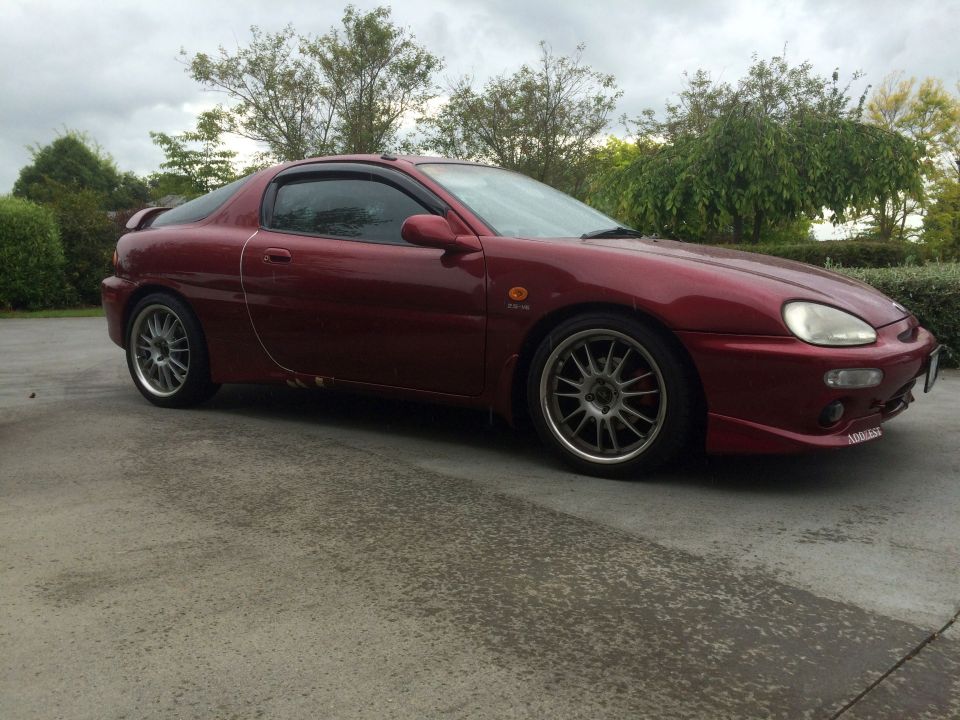 Red Mazda MX-3 coupe parked on wet concrete, side 3/4 front view.