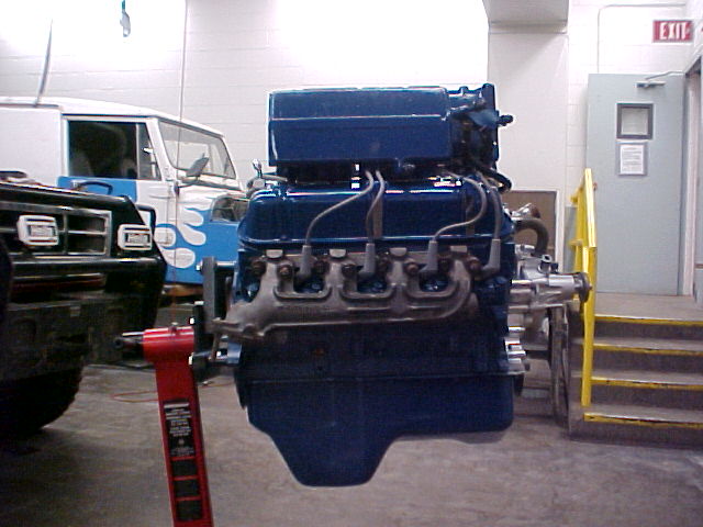 Blue Ford engine on a stand in a shop beside an old flatnose van with flames