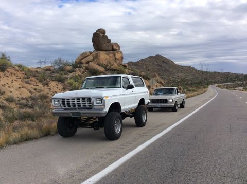 1979 Ford Bronco - A lifted white 1979 Ford Bronco is parked on the side of a desert highway next to a silver pickup truck. The Bronco features large tires an…