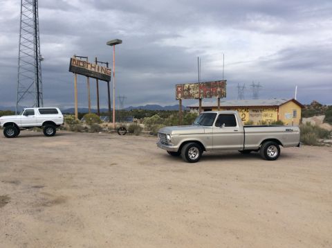 1979 Ford Bronco - A silver 1970s Ford pickup truck is parked on a dirt lot in front of a yellow building with various signs. To the left, a white Ford Bronco…
