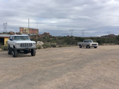 1979 Ford Bronco - A lifted white 1979 Ford Bronco is parked in a dirt lot next to a faded billboard, with a silver pickup truck in the background.