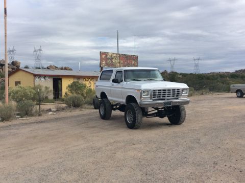 1979 Ford Bronco - A lifted white 1979 Ford Bronco is parked in a dirt lot in front of a dilapidated yellow building with graffiti. The vehicle is modified wi…