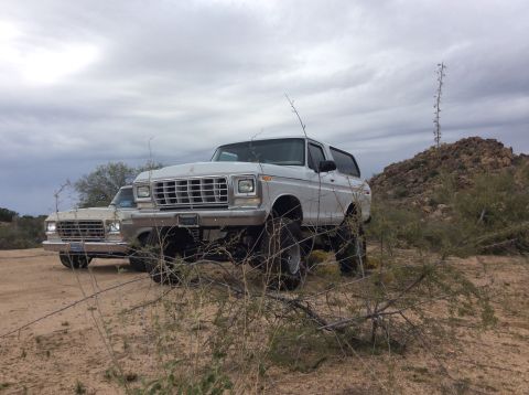 1979 Ford Bronco - Two lifted Ford Broncos are parked in a dry, desert landscape under a cloudy sky. The Bronco in the foreground is white and has large tires…