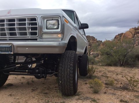 1979 Ford Bronco - A lifted, silver 1979 Ford Bronco is parked in a desert landscape with rocky hills and sparse vegetation under a cloudy sky. The front driv…