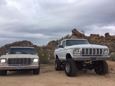 1979 Ford Bronco - Two vintage Ford SUVs are parked in a desert landscape under a cloudy sky. The vehicle on the left is a tan pickup truck, and the vehicle o…