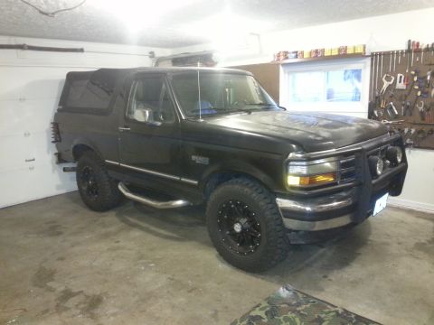 1994 Ford Bronco - A black 1994 Ford Bronco is parked in a garage, facing towards the right. The vehicle has aftermarket wheels and a brush guard on the front.
