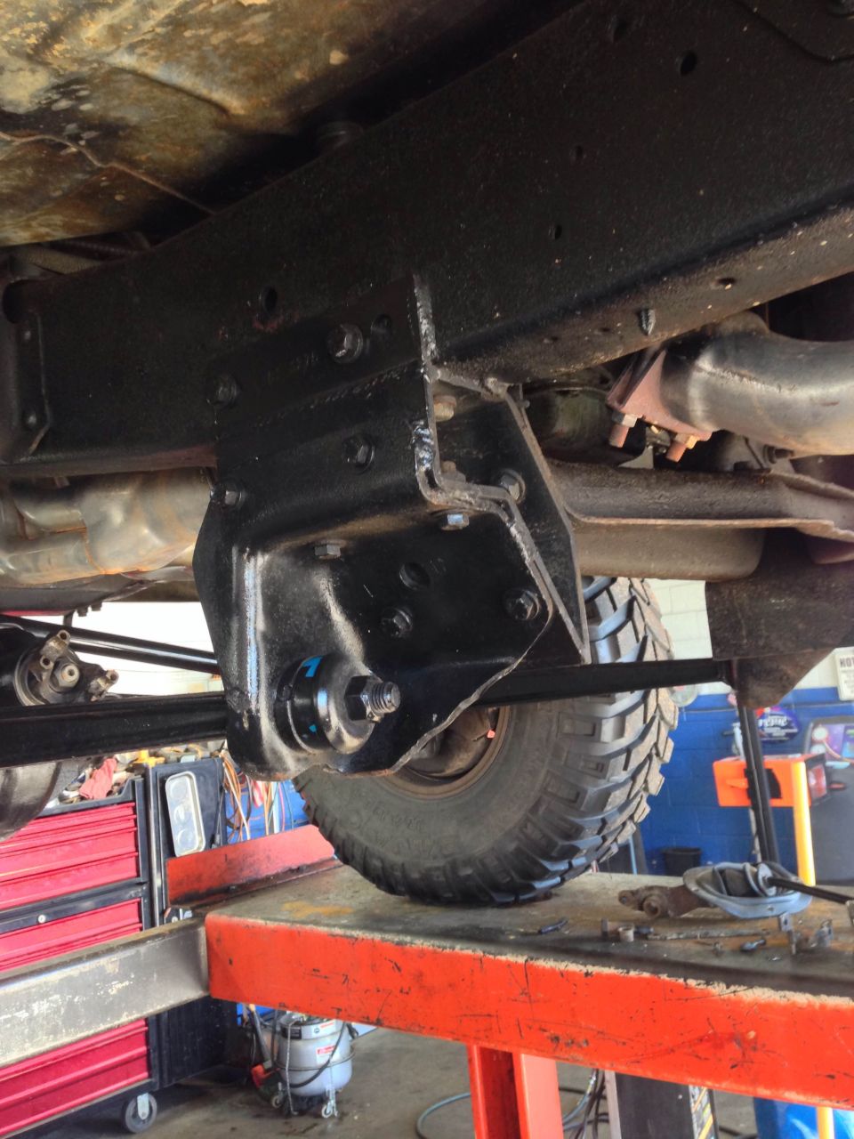 Underside of a black vehicle showing a suspension bracket, tire, and exhaust on an orange lift.