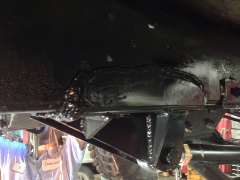 Welded reinforcements on a black vehicle frame, with a welder in the background.