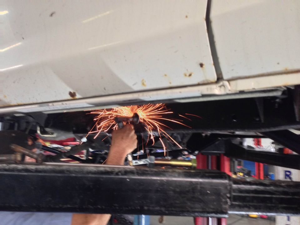 Close-up of sparks flying from an angle grinder cutting metal under a white vehicle on a lift.