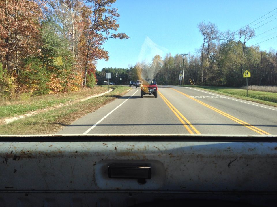 Red 1979 Ford Bronco driving away on a road with autumn trees.