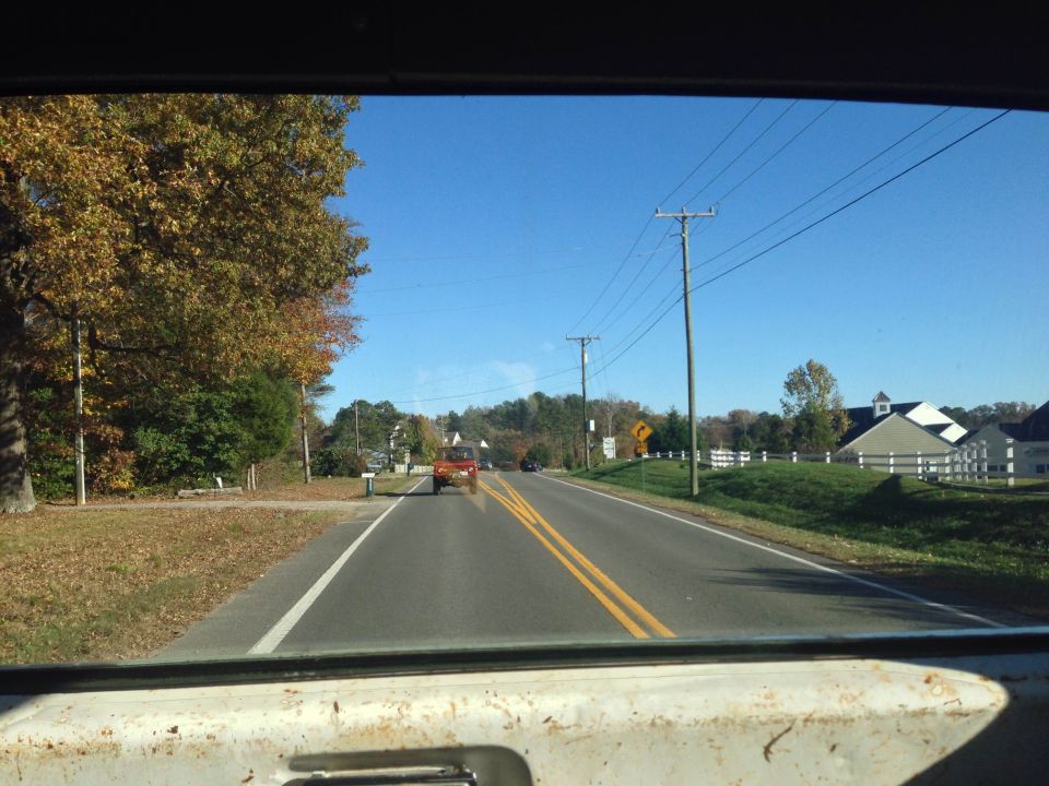 1979 Ford Bronco driving down a road with fall foliage and houses on the side.