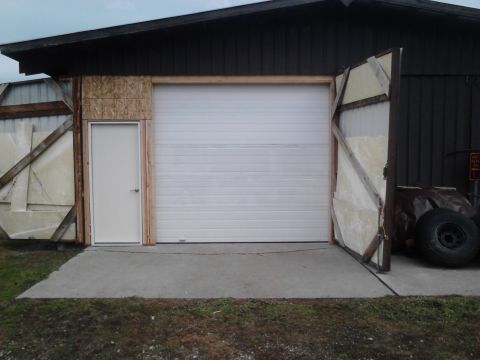 1989 Ford Bronco - A white garage door is partially open, revealing a dark interior. To the left of the garage door is a white pedestrian door. The scene is s…