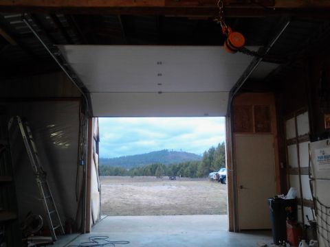 1989 Ford Bronco - View from inside a garage looking out through an open garage door at a field with trees and hills in the background. Several vehicles are p…