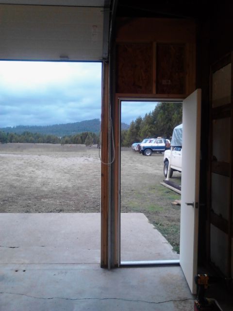 1989 Ford Bronco - View from inside a garage looking out at a cloudy day with a field and trees. Two trucks are parked outside, one white and one blue.