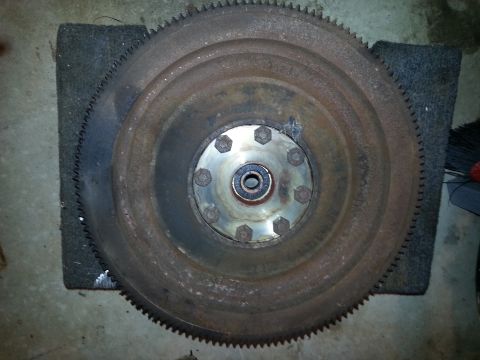 1994 Ford Bronco - A rusty, large metal flywheel with a ring gear is shown resting on a dark gray mat. The flywheel has a central hub with several bolts secur…