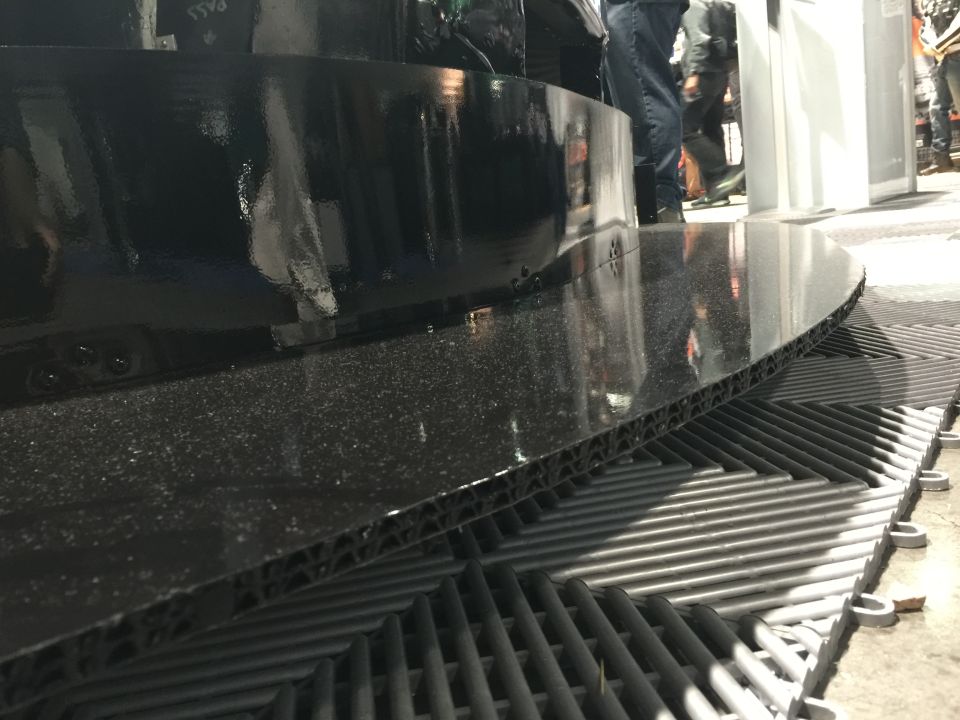 Black display structure with a textured lower section and a glossy curved upper section, resting on a gray grated floor