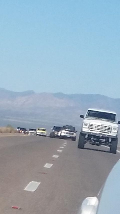 1979 Ford Bronco - A convoy of trucks and SUVs, including a 1979 Ford Bronco, travels down a highway under a clear blue sky.