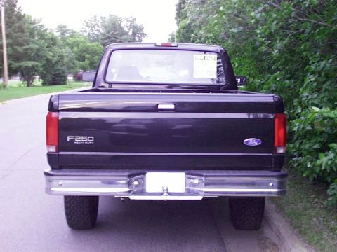1997 Ford F-250 - The rear of a black Ford F-250 Heavy Duty pickup truck is shown, parked curbside with trees and foliage in the background.