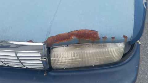 1991 Ford LTD Crown Victoria - Photograph of a blue car's front with a rusted hood edge and headlight, not a document.