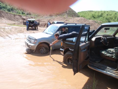1987 Ford Bronco - Two off-road vehicles are stuck in muddy water, with one person leaning out of the driver's side window of the foreground vehicle. The othe…