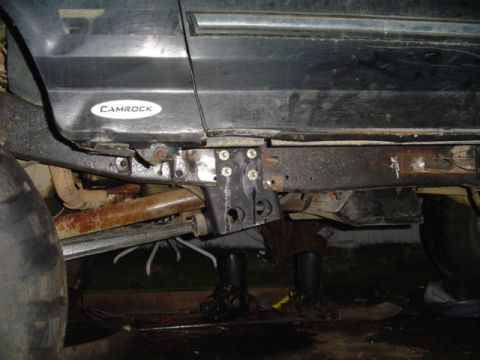 1993 Ford Bronco - Side view of a dark truck's rocker panel and frame rail with a black bracket bolted on with four zinc bolts, the body resting on jack stand…