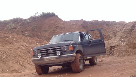 1987 Ford Bronco - A dark colored, older model Ford Bronco is parked on a muddy dirt road in a sandy, hilly area. The driver's side door is open, and the vehi…